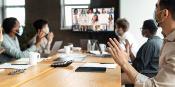 Videoconferencing Concept. Diverse group of workers in face masks making video call with colleagues, sitting at table in boardroom, waving at screen at office, talking on web with remote employees